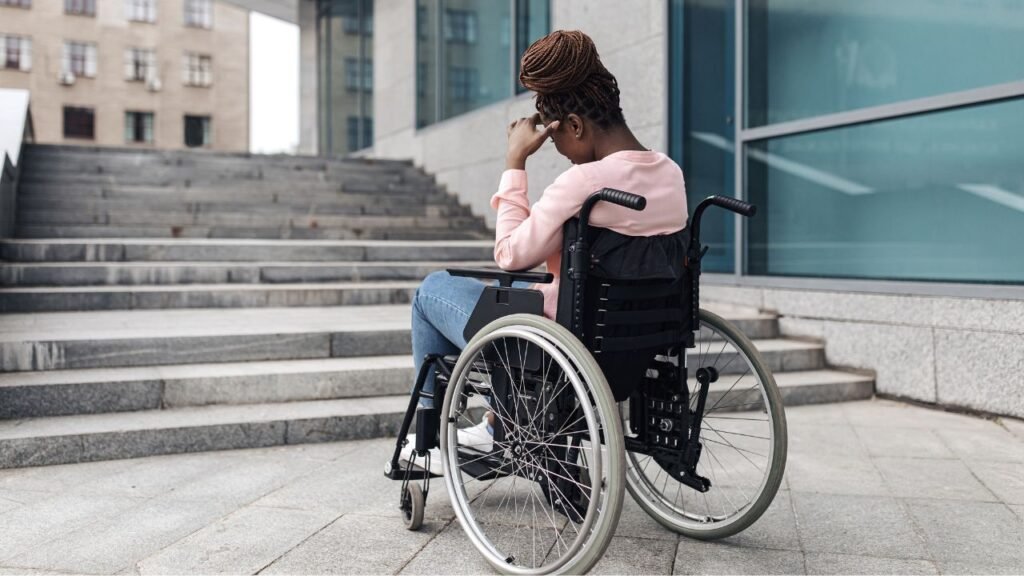 Vemos, de costas, uma mulher negra, cadeirante, vestindo blusa de malha manga longa rosa e calça jeans azul claro e tênis branco. Ela está diante de uma escadaria na entrada de um prédio. Coloca a mão na testa em sinal de frustração. A cena representa a barreira de acessibilidade ainda presente em muitos locais públicos.