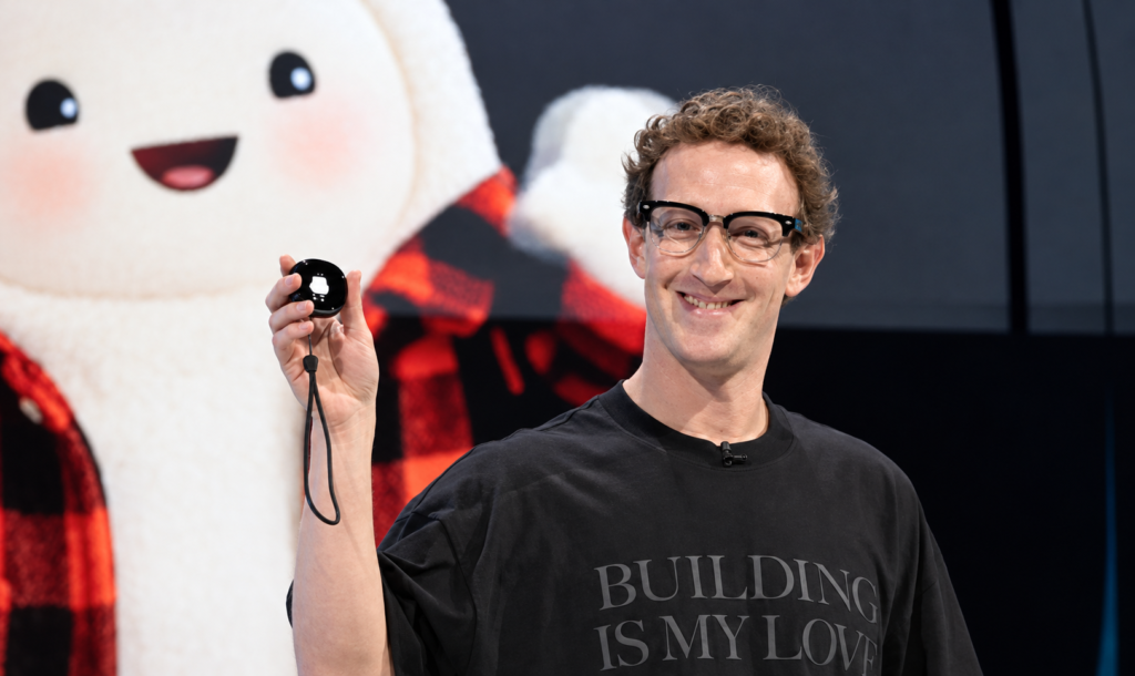 Mark Zuckerberg sorri, usando óculos de armação preta e camiseta preta. Com a mão direita levantada, segura um pequeno dispositivo preto arredondado, com uma alça pendurada. Ao fundo, aparece um grande personagem branco sorridente, vestido com camisa xadrez vermelha e preta.